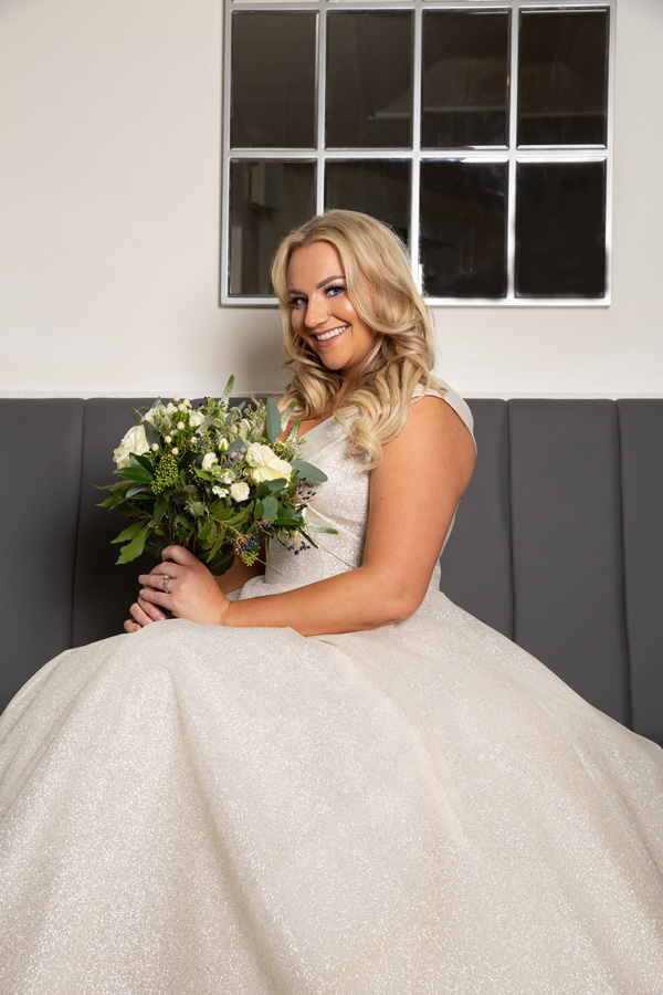 Model sitting on a grey bench with a floral bouquet wearing Alice Bridal Dress stocked by Roberta's Bridal, Burslem