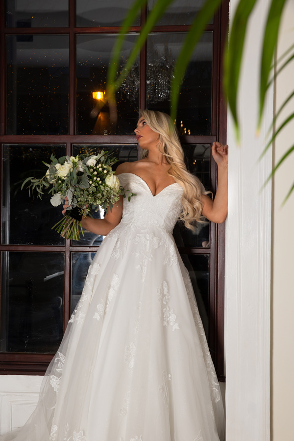 Model standing in a doorway with a bouquets wearing Alice Bridal Dress stocked by Roberta's Bridal, Burslem