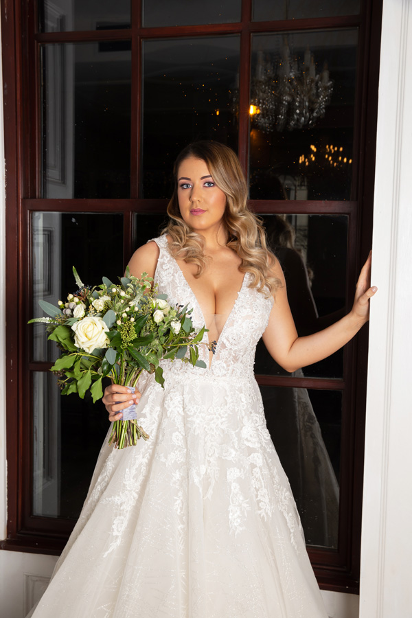 Model leaning against a doorway with a bouquets wearing Alice Bridal Dress stocked by Roberta's Bridal, Burslem